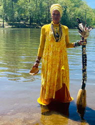 Image of Iya FaBambi Shangodeye standing in a river with trees behind her. She is wearing an orange long dress and head piece and holding a staff.