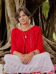 Picture of Rebecca Owen wearing a read shirt and white skirt sitting cross legged in front of a tree trunk.